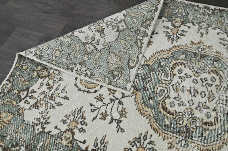 Op de afbeelding: Een vintage tapijt met een witte achtergrond en een groen, bruin en beige bloemenpatroon.