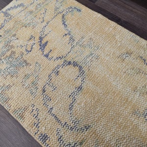 K&ouml;nnte beinhalten: Ein hellgelber Teppich mit einem blauen und gr&uuml;nen abstrakten Muster. Der Teppich hat eine gewebte Textur.