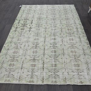 May include: A rectangular, light green rug with a repeating floral and geometric pattern in shades of gray and brown. The rug is placed on a dark wood floor, showcasing its intricate design and texture. The rug's edges are finished with a subtle border.