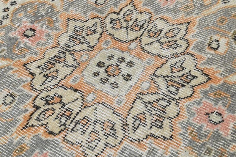 Puede incluir: Primer plano de una alfombra vintage con un dise&ntilde;o de medall&oacute;n central. La alfombra presenta una paleta de colores apagados de gris, beige y melocot&oacute;n, con intrincados patrones florales y geom&eacute;tricos. La textura de la alfombra parece suave y desgastada, lo que sugiere edad y uso.