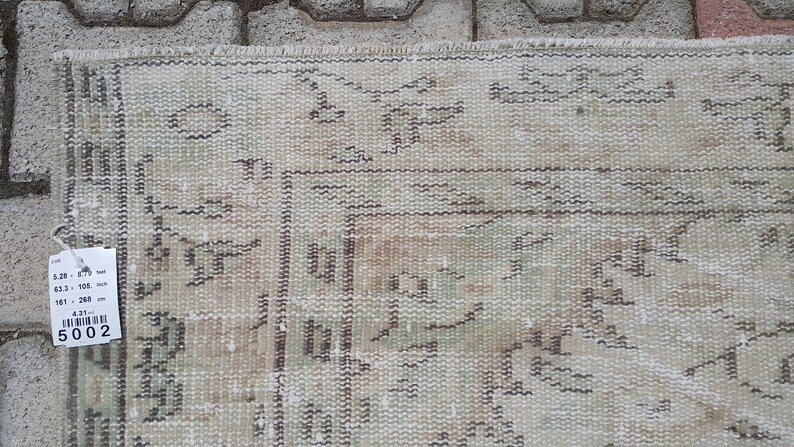May include: A beige rug with a geometric pattern in brown and green. The rug is 63.5 inches by 101 inches, or 161 cm by 256 cm. The rug has a tag with the number 5002 on it.