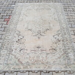 May include: A vintage cream-colored rug with a floral pattern in shades of brown. The rug has a distressed look and is laid out on a gray brick surface.