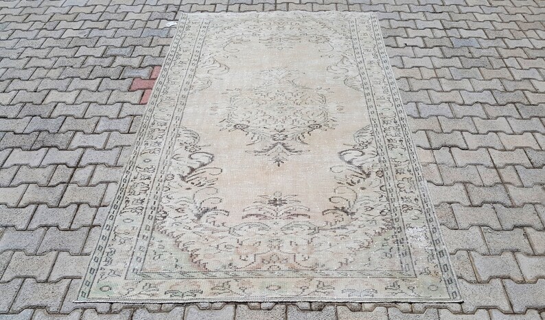 May include: A vintage cream-colored rug with a floral pattern in shades of brown. The rug has a distressed look and is laid out on a gray brick surface.