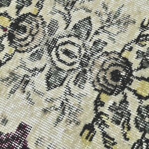 May include: Close-up of a beige rug with a floral pattern in shades of black, gray, and yellow. The rug has a distressed, vintage look.