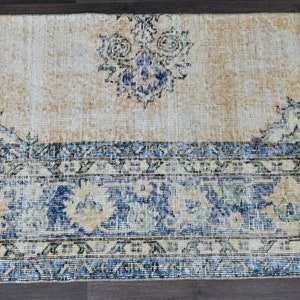 Op de afbeelding: Een vintage tapijt met een cr&egrave;mekleurige achtergrond en blauwe en beige bloemmotieven. Het tapijt heeft een verweerde uitstraling en een franje rand.