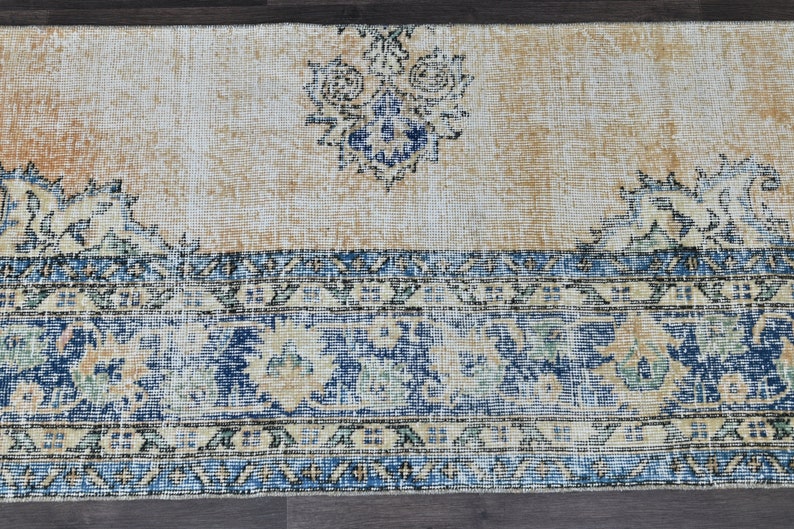 Op de afbeelding: Een vintage tapijt met een cr&egrave;mekleurige achtergrond en blauwe en beige bloemmotieven. Het tapijt heeft een verweerde uitstraling en een franje rand.