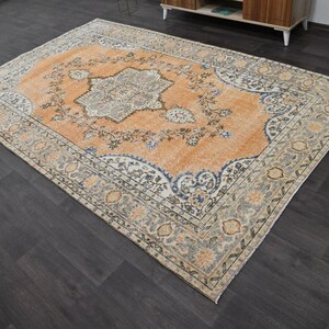 Puede incluir: Una alfombra rectangular antigua con un intrincado dise&ntilde;o floral. La alfombra presenta un medall&oacute;n central en tonos naranja, azul y gris, rodeado por un borde con colores similares. Est&aacute; colocada sobre un suelo de madera oscura.