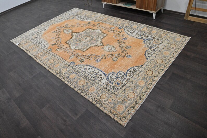 Puede incluir: Una alfombra rectangular antigua con un intrincado dise&ntilde;o floral. La alfombra presenta un medall&oacute;n central en tonos naranja, azul y gris, rodeado por un borde con colores similares. Est&aacute; colocada sobre un suelo de madera oscura.