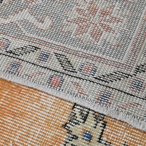 Puede incluir: Primer plano de una secci&oacute;n de una alfombra vintage con un patr&oacute;n detallado. La alfombra presenta un borde gris claro con un dise&ntilde;o geom&eacute;trico repetitivo en negro, azul y rosa. El &aacute;rea central es de color naranja c&aacute;lido con un patr&oacute;n de rayas.