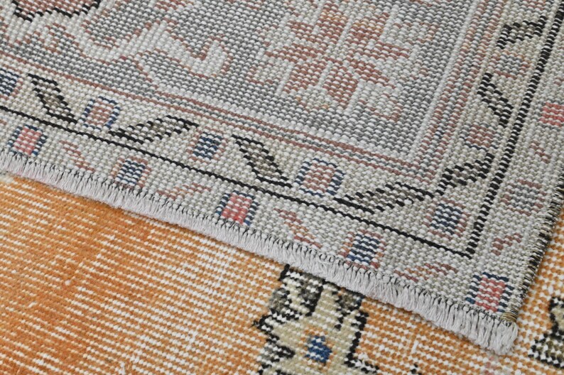 Puede incluir: Primer plano de una secci&oacute;n de una alfombra vintage con un patr&oacute;n detallado. La alfombra presenta un borde gris claro con un dise&ntilde;o geom&eacute;trico repetitivo en negro, azul y rosa. El &aacute;rea central es de color naranja c&aacute;lido con un patr&oacute;n de rayas.