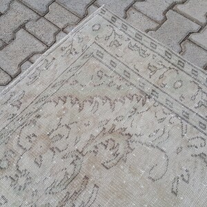 May include: A close-up of a cream-colored rug with a faded, intricate pattern in a darker shade of cream. The rug is lying on a gray brick surface.