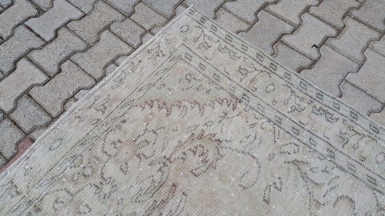 May include: A close-up of a cream-colored rug with a faded, intricate pattern in a darker shade of cream. The rug is lying on a gray brick surface.