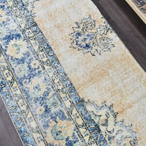Op de afbeelding: Een vintage tapijt met een blauw en cr&egrave;me patroon. Het tapijt heeft een vervaagde uitstraling en een verweerde textuur. Het tapijt ligt op een donkere houten vloer.
