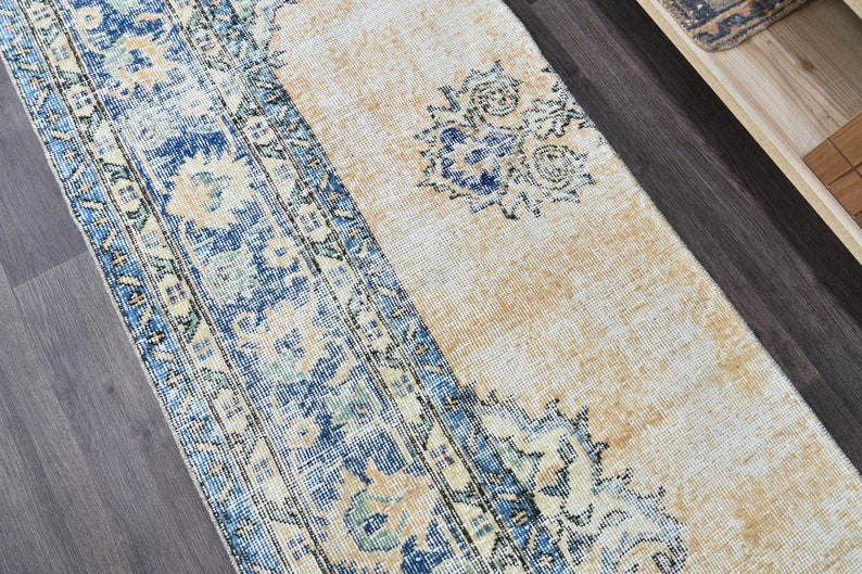 Op de afbeelding: Een vintage tapijt met een blauw en cr&egrave;me patroon. Het tapijt heeft een vervaagde uitstraling en een verweerde textuur. Het tapijt ligt op een donkere houten vloer.