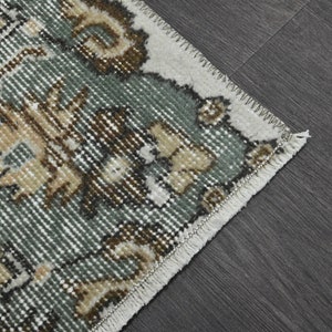 Op de afbeelding: Close-up van een tapijt met een groen, bruin en wit bloemenpatroon. Het tapijt heeft een witte franje rand.