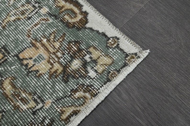Op de afbeelding: Close-up van een tapijt met een groen, bruin en wit bloemenpatroon. Het tapijt heeft een witte franje rand.