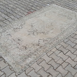 May include: A vintage rug with a floral pattern in shades of beige and brown. The rug is laid out on a gray brick surface.