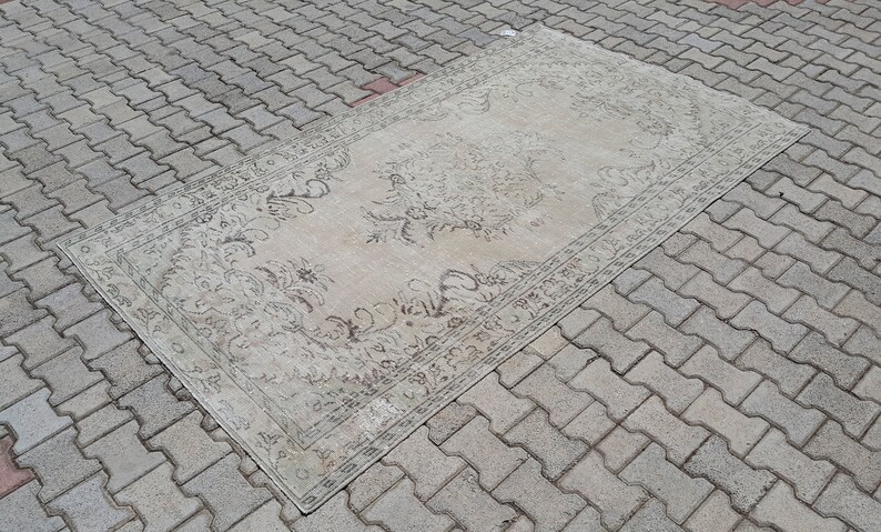 May include: A vintage rug with a floral pattern in shades of beige and brown. The rug is laid out on a gray brick surface.