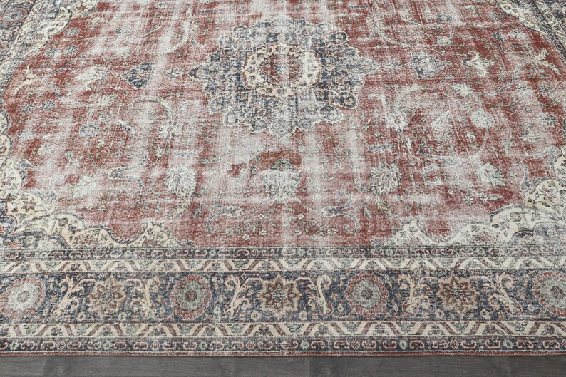 Pode incluir: Um tapete de estilo vintage com um fundo vermelho desbotado e padr&otilde;es florais intrincados em tons de creme, azul e cinza. O tapete tem um visual desgastado e uma borda com um design geom&eacute;trico.