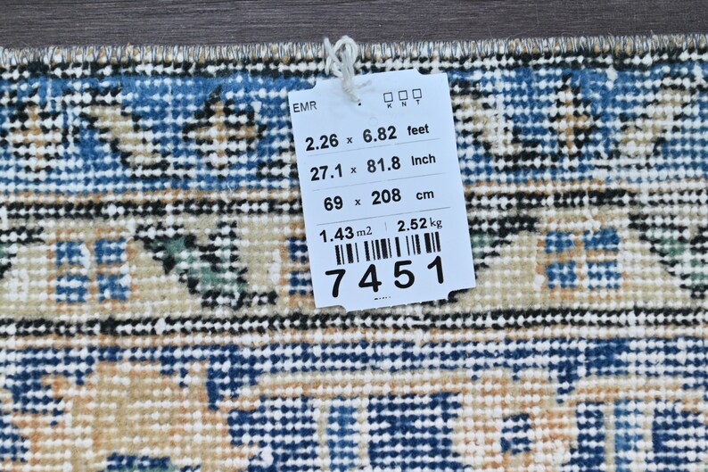 Op de afbeelding: Een wit label met zwarte tekst met de afmetingen van een tapijt in voet, inch en centimeter. Het label geeft ook de oppervlakte van het tapijt in vierkante meter en het gewicht in kilogram weer. Het label heeft een barcode en het nummer 7451 staat erop gedrukt.