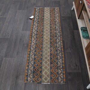 Può includere: Un tappeto rettangolare a motivi con una striscia centrale beige affiancata da disegni intricati in arancione, grigio e bianco. I bordi del tappeto presentano un motivo geometrico ripetuto. Il tappeto è posizionato su un pavimento in legno scuro.