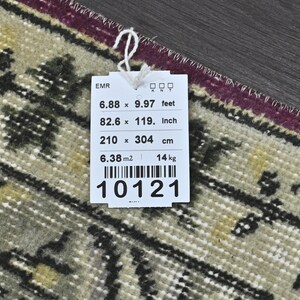 May include: A white tag with black text and a barcode. The tag lists the dimensions of a rug in feet, inches, and centimeters. The rug is 6.88 feet by 9.97 feet, 82.6 inches by 119 inches, and 210 centimeters by 304 centimeters. The tag also lists the rug's area as 6.38 square meters and its weight as 14 kilograms. The tag number is 10121.