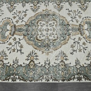Op de afbeelding: Een vintage tapijt met een cr&egrave;mekleurige achtergrond en een centraal medaillon in tinten groen, bruin en beige. Het tapijt heeft een herhalend bloemenpatroon en ingewikkelde details.