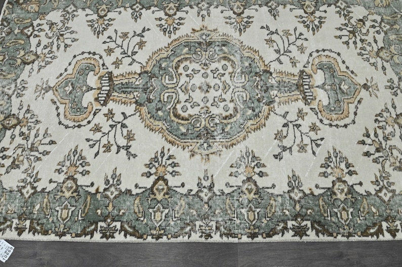 Op de afbeelding: Een vintage tapijt met een cr&egrave;mekleurige achtergrond en een centraal medaillon in tinten groen, bruin en beige. Het tapijt heeft een herhalend bloemenpatroon en ingewikkelde details.