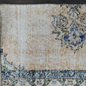 Op de afbeelding: Een beige kleed met een blauw en wit bloemenpatroon. Het kleed heeft een franje rand en is gemaakt van geweven materiaal.