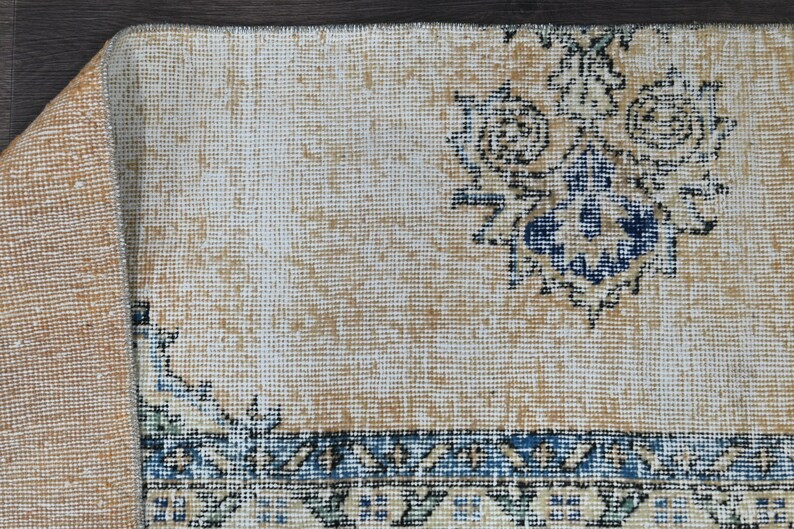 Op de afbeelding: Een beige kleed met een blauw en wit bloemenpatroon. Het kleed heeft een franje rand en is gemaakt van geweven materiaal.