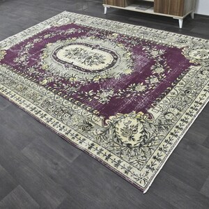 May include: A vintage rug with a floral pattern in shades of purple, cream, and black. The rug has a distressed look and a fringed edge.