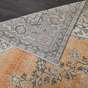 Puede incluir: Una alfombra de estilo antiguo con un aspecto desgastado y descolorido. La alfombra presenta un campo central naranja c&aacute;lido con patrones florales y geom&eacute;tricos en negro, crema y gris. El borde es gris con dise&ntilde;os intrincados. La alfombra est&aacute; parcialmente doblada.