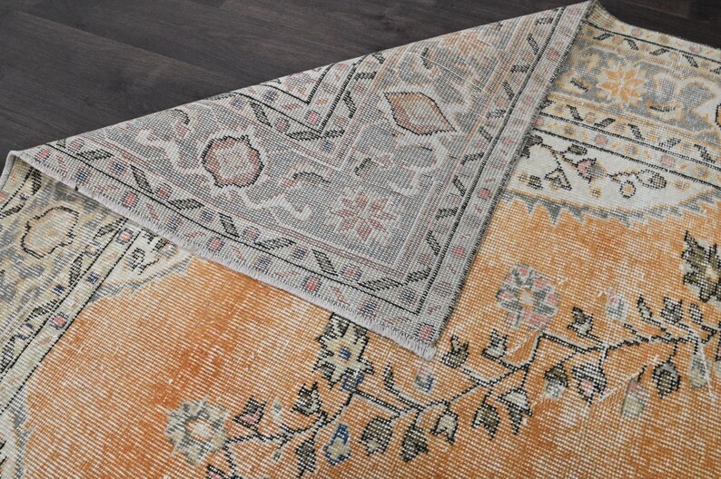 Puede incluir: Una alfombra de estilo antiguo con un aspecto desgastado y descolorido. La alfombra presenta un campo central naranja c&aacute;lido con patrones florales y geom&eacute;tricos en negro, crema y gris. El borde es gris con dise&ntilde;os intrincados. La alfombra est&aacute; parcialmente doblada.