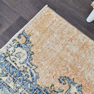 Op de afbeelding: Een vintage tapijt met een verbleekte oranje achtergrond en een blauw en wit bloemenpatroon. Het tapijt heeft een verweerde uitstraling en is gemaakt van een zacht, geweven materiaal.