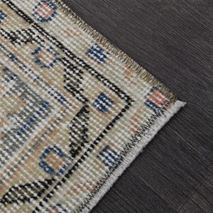 Puede incluir: Primer plano de una secci&oacute;n de una alfombra vintage con un patr&oacute;n geom&eacute;trico. La alfombra presenta un fondo crema con detalles azules, negros y naranjas. Los bordes est&aacute;n deshilachados y la alfombra est&aacute; colocada sobre una superficie de madera oscura.