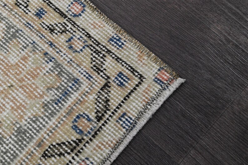 Puede incluir: Primer plano de una secci&oacute;n de una alfombra vintage con un patr&oacute;n geom&eacute;trico. La alfombra presenta un fondo crema con detalles azules, negros y naranjas. Los bordes est&aacute;n deshilachados y la alfombra est&aacute; colocada sobre una superficie de madera oscura.