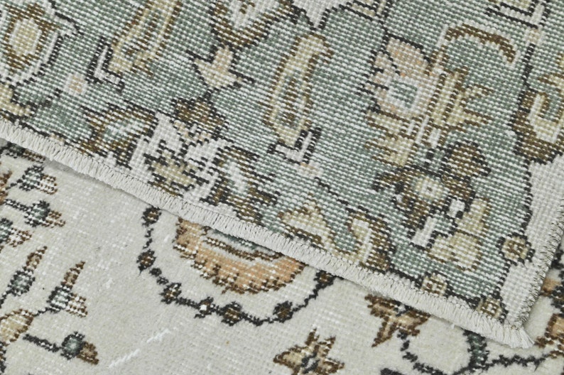 Op de afbeelding: Close-up van een tapijt met een gedetailleerd bloemenpatroon in beige, bruin en groene tinten. Het tapijt heeft een witte franje rand.