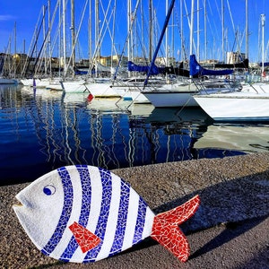 Może przedstawiać: Rzeźba ryby z białej i niebieskiej mozaiki z czerwonymi akcentami. Ryba stoi na szarej powierzchni przed portem z żaglówkami.