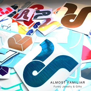 May include: A collection of colorful, abstract stickers with a variety of shapes and designs. The stickers are arranged in a random pattern and feature a blue, teal, gold, red, and white color scheme. The stickers are labeled "Almost Familiar" and "Funky Jewelry & Gifts".