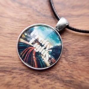 May include: A round pendant necklace with the words "Pretty Lights" in white script. The pendant features a colorful image of lights and a sky. The necklace has a silver-colored bail and a black cord.