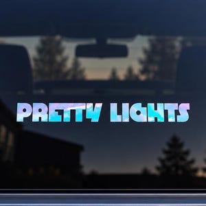 May include: A vinyl decal applied to a vehicle rear window featuring the text PRETTY LIGHTS in a bold, sans-serif font. The lettering displays a holographic, iridescent color pattern that shifts between shades of blue, purple, and white.
