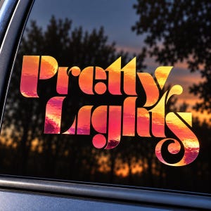 May include: A vinyl decal applied to a car window featuring the text Pretty Lights in a bold, retro-inspired font. The lettering is filled with a vibrant sunset gradient pattern, transitioning from deep orange and red to bright yellow hues.