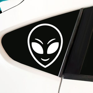 May include: White alien face sticker on a black background. The sticker is on a car window.
