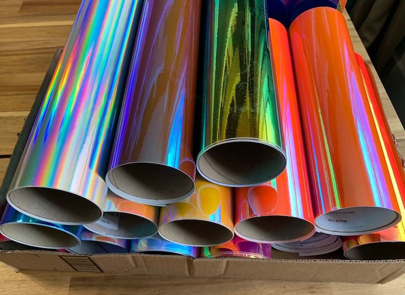 Peut inclure: Un ensemble de rouleaux de vinyle iridescent de diff&eacute;rentes couleurs, notamment argent, or, orange et bleu. Les rouleaux sont empil&eacute;s dans une bo&icirc;te en carton.