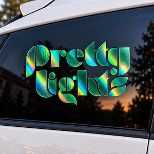 May include: A holographic decal applied to a car window featuring the text "pretty lights" in a stylized, bold, sans-serif font. The material reflects a spectrum of colors including green, yellow, and blue, creating a vibrant, shifting effect.