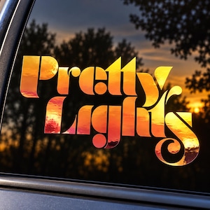 May include: A vinyl decal applied to a car window featuring the text Pretty Lights in a bold, stylized font. The letters are filled with a vibrant sunset gradient of orange, yellow, and deep red, reflecting the colors of the sky visible through the glass.