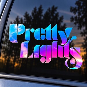 May include: A vinyl decal sticker applied to a car window featuring the text Pretty Lights in a stylized, bold font. The lettering displays a vibrant, holographic gradient effect that shifts between shades of blue, purple, and bright pink.
