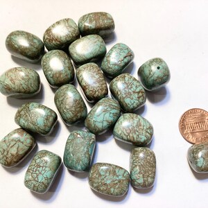 May include: A collection of turquoise-colored, polished stones with brown veining. The stones are oval-shaped and vary in size. A penny is included for size reference. These stones are likely used for jewelry making or crafts.