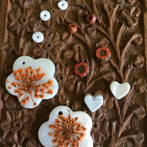 May include: Two white shell flower charms with orange flower details, two white heart charms, and five white and red beads on a brown wooden background.