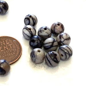 May include: A collection of round, striped beads in shades of gray, brown, and blue, with a hole drilled through each. A U.S. one-cent coin is included for size reference. The beads have a polished finish and are likely for jewelry making.